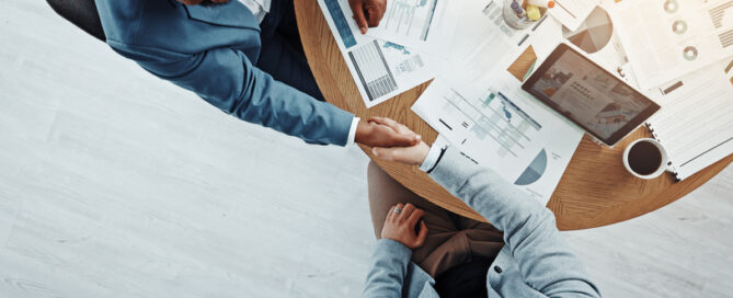 Two men in a business meeting shaking hands with papers on table.